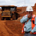 A dump truck drives through piles of red-coloured ore towards two workers in the foreground