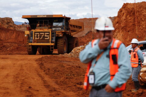 A dump truck drives through piles of red-coloured ore towards two workers in the foreground